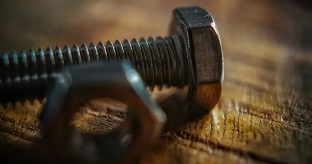 A shallow depth of field, macro photograph of a steel hex bolt and a matching hexagonal nut resting on a textured wooden surface. The bolt head and sharp threads are in focus on the right, while the nut in the foreground is blurred.