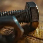 A shallow depth of field, macro photograph of a steel hex bolt and a matching hexagonal nut resting on a textured wooden surface. The bolt head and sharp threads are in focus on the right, while the nut in the foreground is blurred.