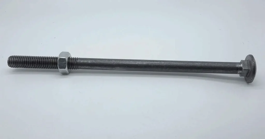 A single, elongated galvanized metal carriage bolt with a round head, a square neck, and a mostly threaded shank, positioned horizontally against a plain white studio background. A single hexagonal nut is threaded partially onto the shank, showing the contrasting light-colored metal of the nut against the dark grey bolt.