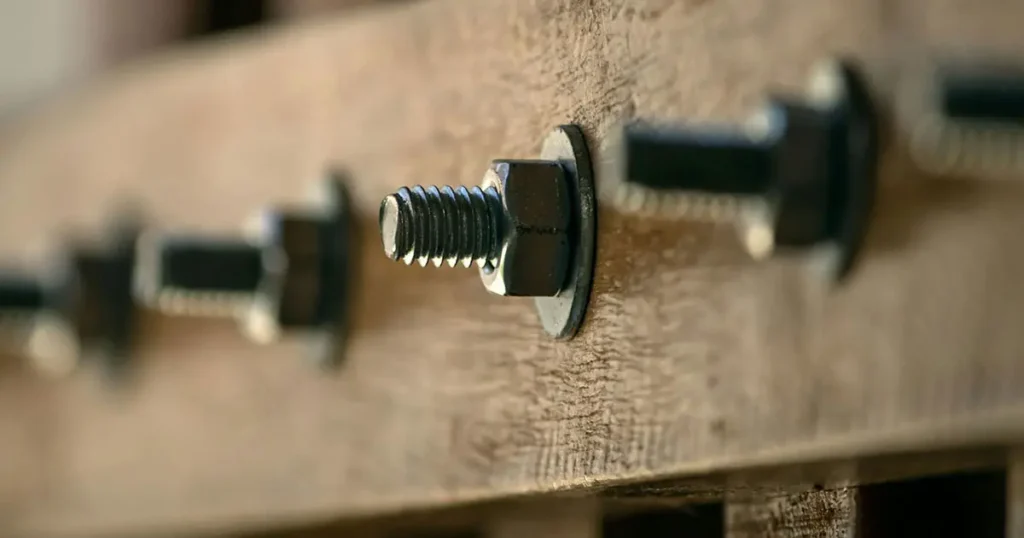 A close-up photograph of a timber beam connection, showing multiple metal carriage bolt assemblies holding wooden elements together. The central bolt features a threaded end, a hexagonal nut, and a flat washer, all tightly fastened. Other similar bolt assemblies are visible to the left and right, receding into a shallow depth of field. The texture of the raw wood is prominent.