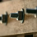 A close-up photograph of a timber beam connection, showing multiple metal carriage bolt assemblies holding wooden elements together. The central bolt features a threaded end, a hexagonal nut, and a flat washer, all tightly fastened. Other similar bolt assemblies are visible to the left and right, receding into a shallow depth of field. The texture of the raw wood is prominent.