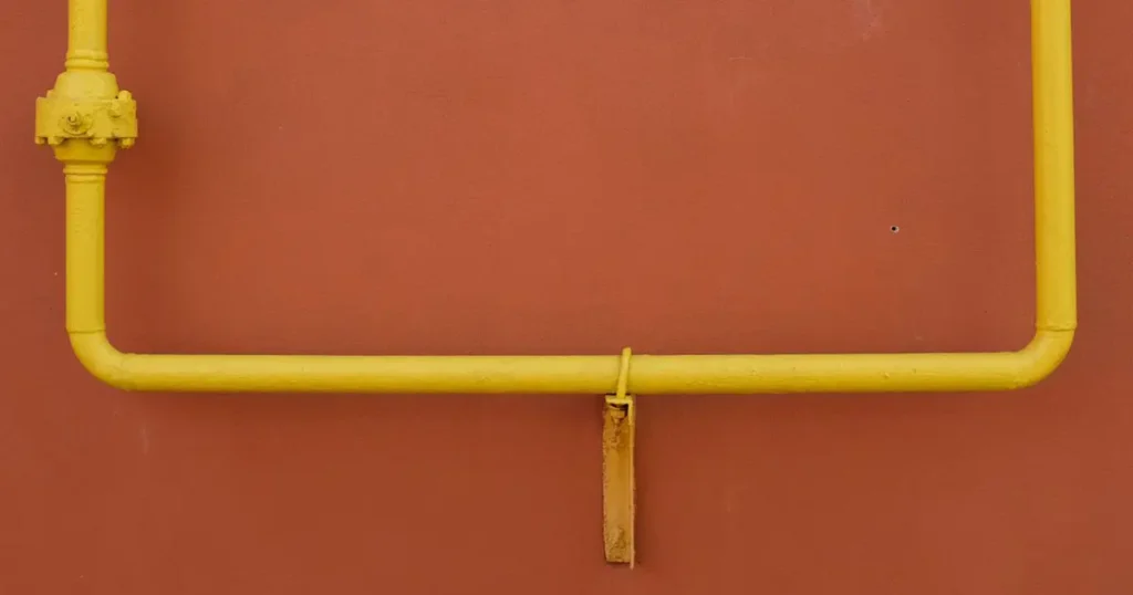 Close-up detail of a painted yellow U-bolt clamp securing a horizontal gas pipeline section to a metal support bracket on a terracotta-colored industrial wall
